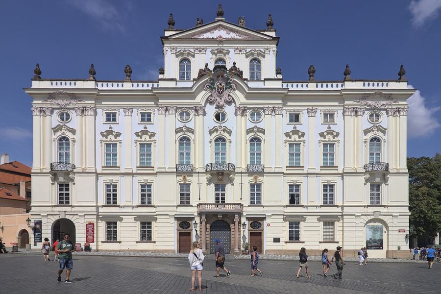 A wide cream rococo palace facade with pilasters, a coat of arms above the central balcony and ornamental gables, tourists crossing the cobbled square in front.
