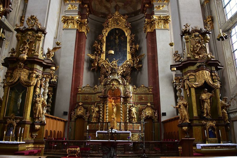 Baroque church interior looking towards a tall gilded high altar with a dark painting, flanked by two side altars with twisted columns and gilt statues.