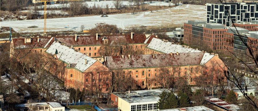 Aerial view of a large pink baroque block with red tile roofs arranged round courtyards, bare winter trees around it and modern glass and brick office buildings behind.