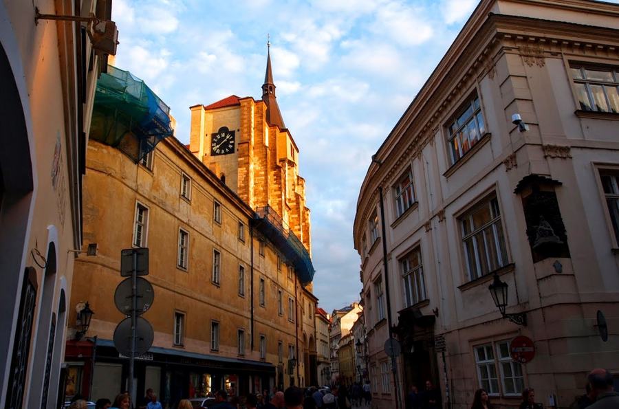 A Gothic church tower with a clock face and slender spire rising in evening sunlight above the roofs of a narrow street, people walking below.