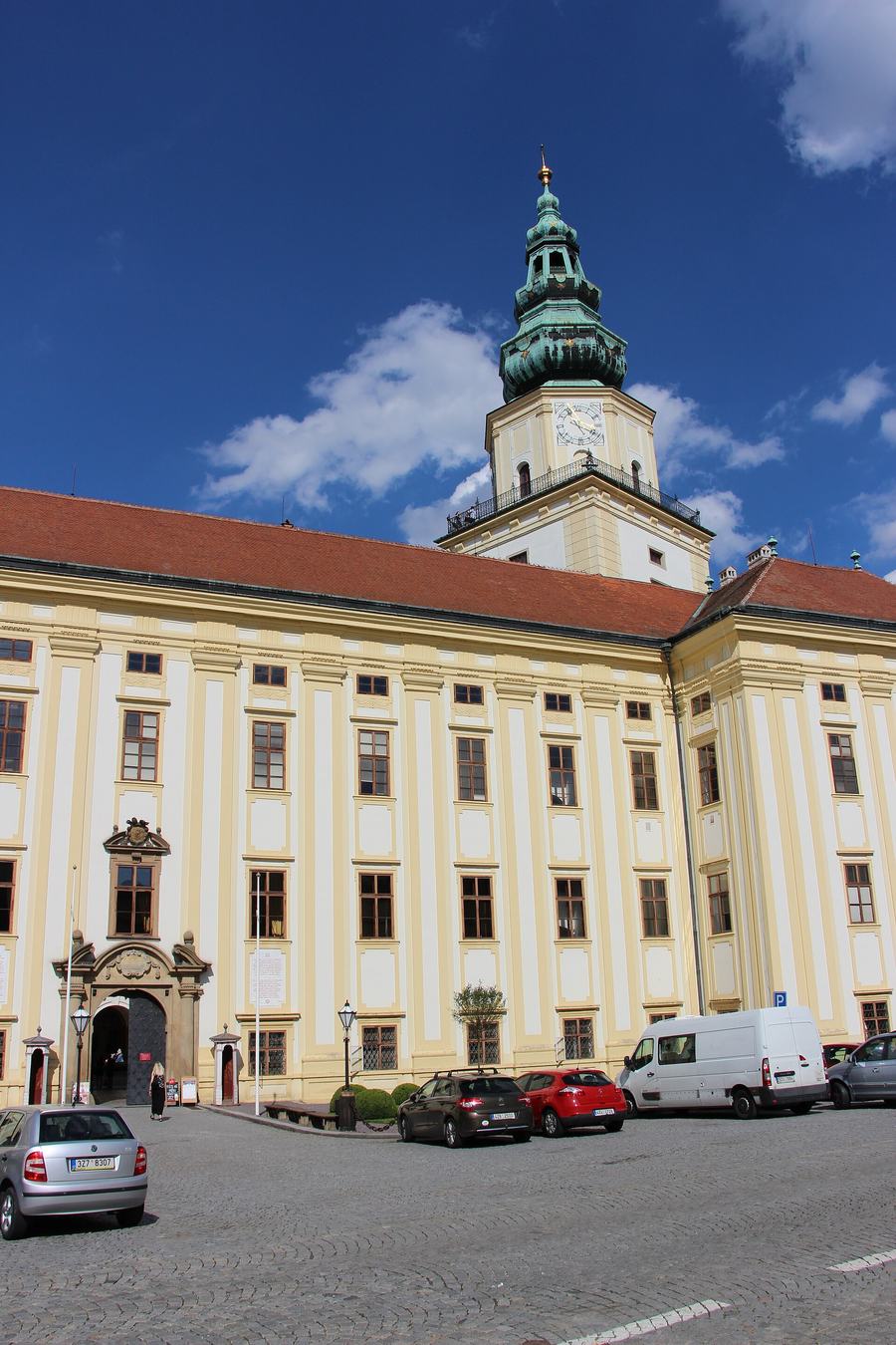 A pale yellow baroque palace wing with pilasters and a red roof, a tall clock tower with a green onion dome behind it, cars parked on the cobbles in front.