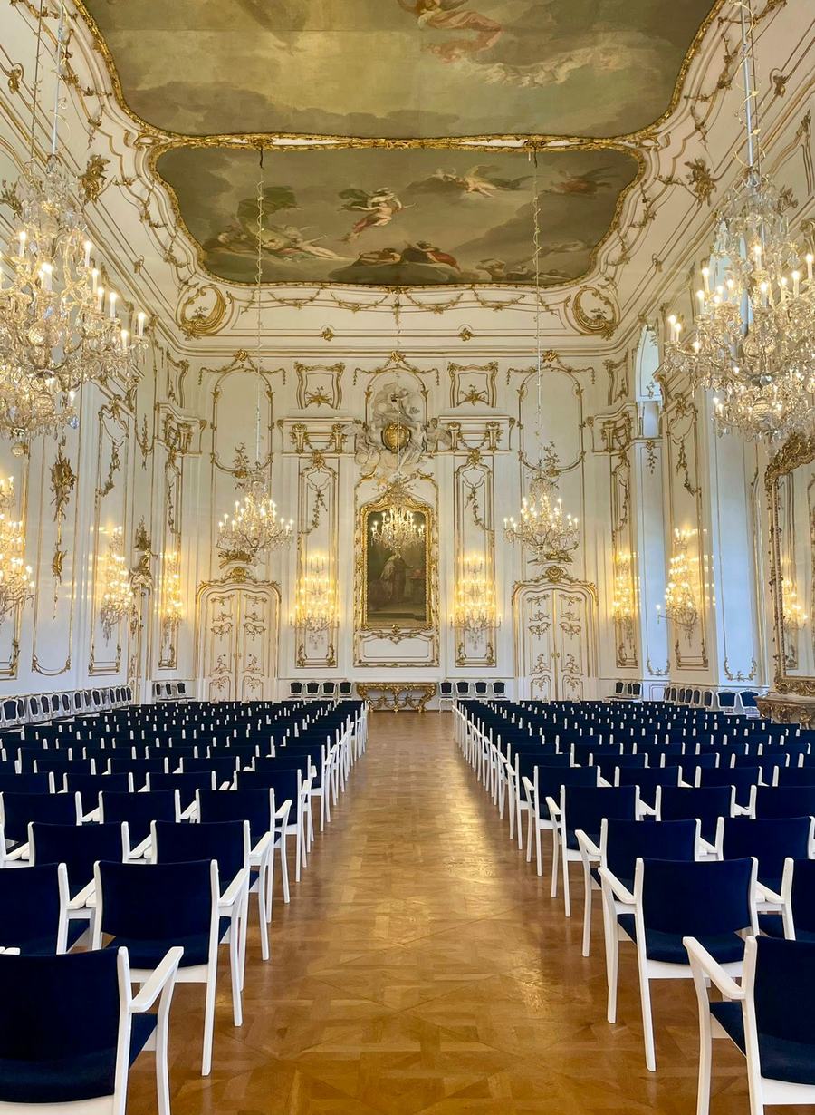 A long white and gold rococo hall with a painted ceiling, crystal chandeliers and wall sconces, rows of blue chairs set out on a parquet floor facing a painting at the far end.