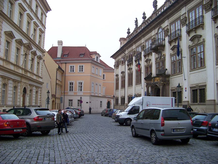 A cobbled square lined with palaces, rows of cars parked down both sides, a delivery van in the middle distance and two people walking on the cobbles.