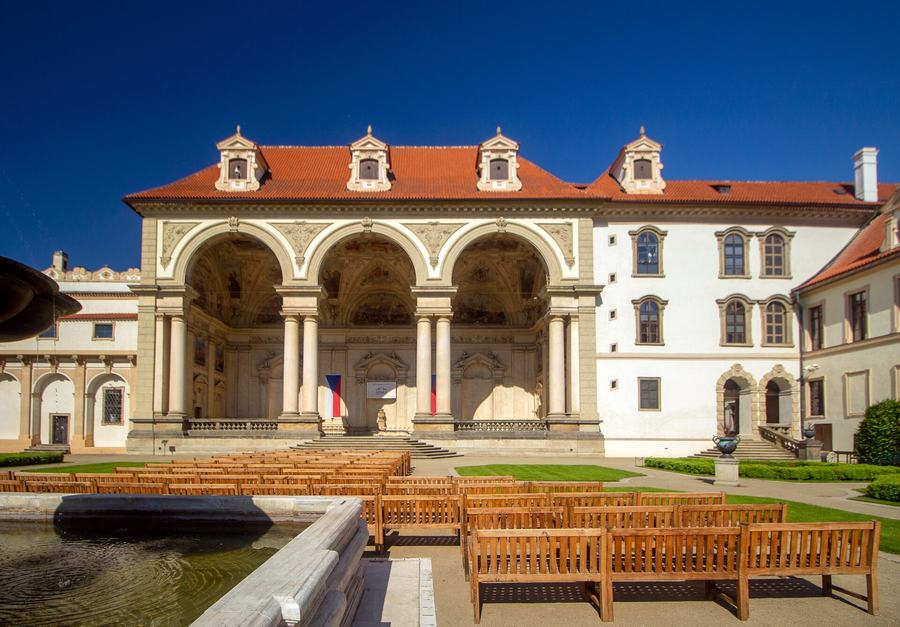 A three-arched Renaissance loggia with frescoed vaults facing a lawn, rows of wooden benches in front of it, a fountain basin at the left and Czech flags hanging in the arches.
