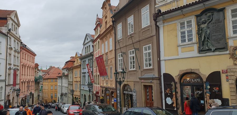 A steep street of tall baroque and renaissance houses with shop fronts, hanging signs and banners, crowded with people and parked cars, a relief plaque of Jan Neruda on the right.