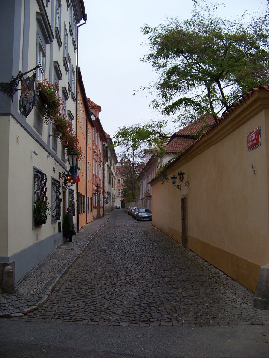 A narrow cobbled lane between a row of pastel houses with window boxes on the left and a long ochre garden wall on the right, cars parked at the far end.