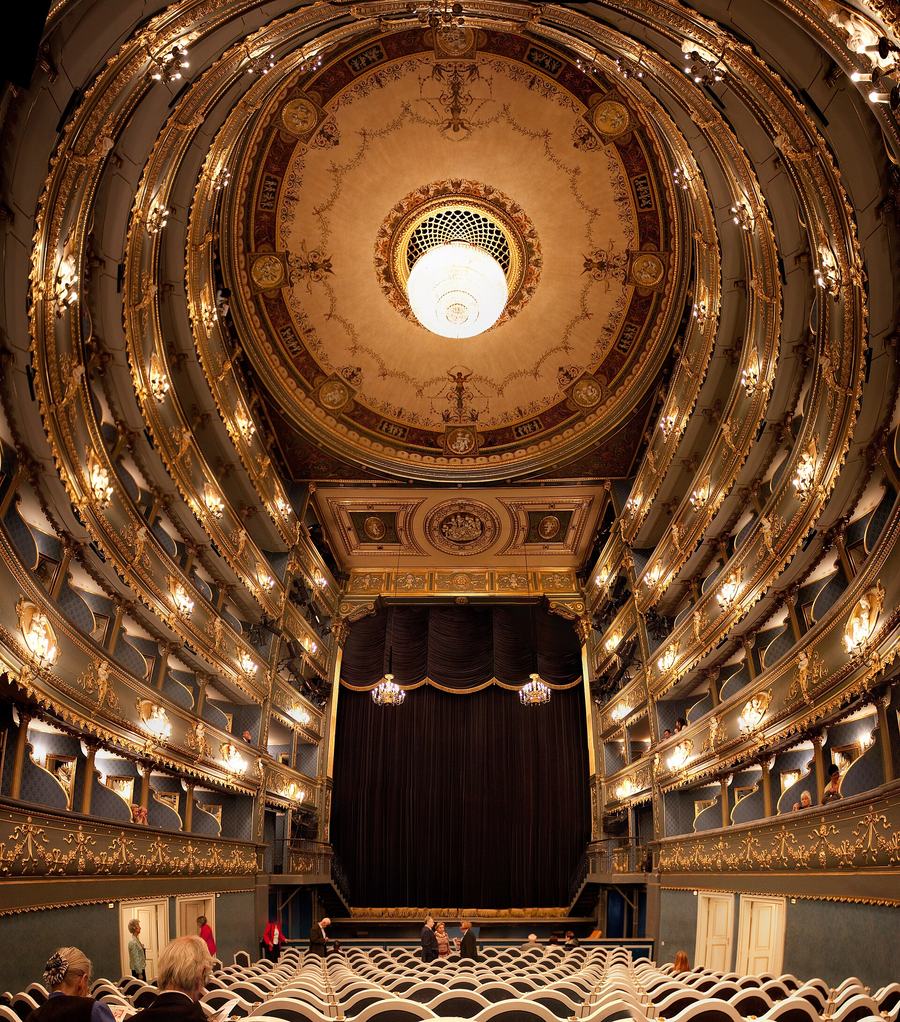 Interior of a small classical theatre seen from the last row: four tiers of boxes curving round to a dark stage curtain, blue panels and heavy gilt decoration, a painted ceiling with a central chandelier.