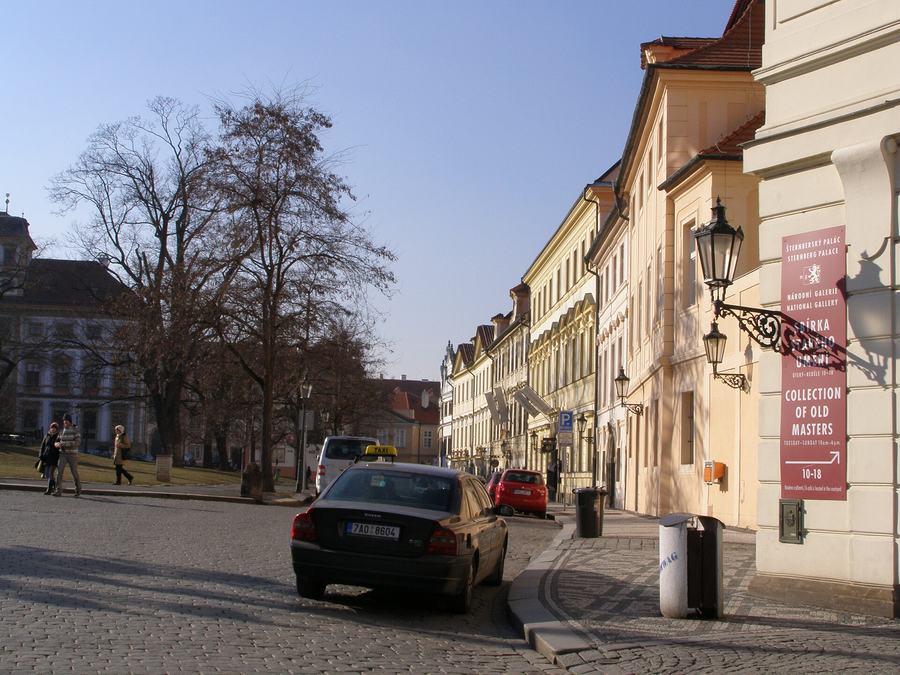 A broad cobbled square sloping away, bare trees and grass on the left, a row of pale palaces on the right with a National Gallery banner, a taxi parked at the kerb.