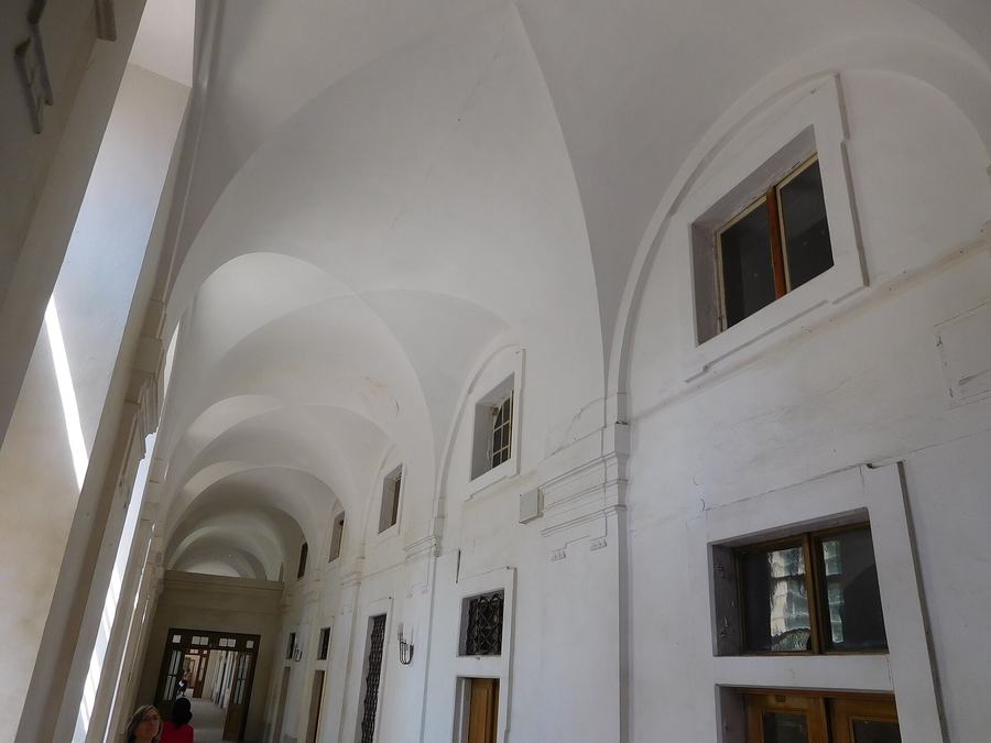 A long whitewashed corridor with a row of low barrel vaults, small square windows high in the wall, a wooden door at the far end and two people standing in it.