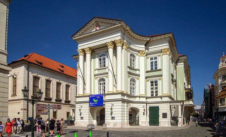 A pale green and white classical theatre with a columned portico and pediment standing on a cobbled square, a banner hanging under the portico, people walking past.