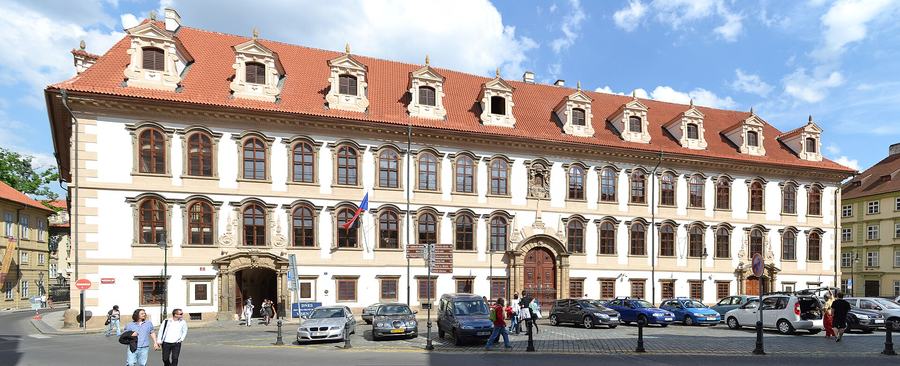 A long white palace facade of three window storeys under a red mansard roof with dormers, two stone portals, cars parked along the cobbled square in front.
