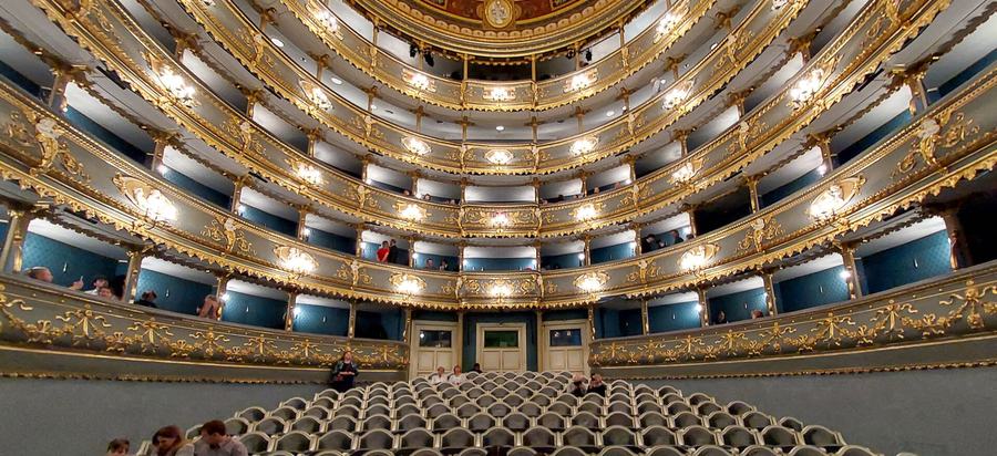View from a stage towards curved tiers of theatre boxes in blue and gold, rows of empty stalls seats below, a few people sitting in the boxes.