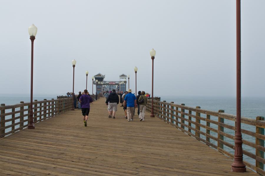 Wooden deck of Oceanside Pier lined with lamp posts, people walking toward the building at the far end