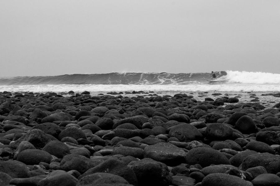 Black-and-white view of a cobblestone beach at Trestles, San Onofre State Beach, with a surfer riding a wave