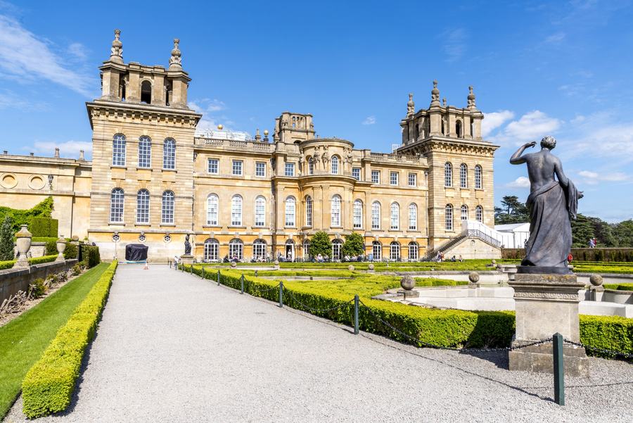 A huge baroque palace of golden stone with towers and a colonnaded centre, seen along a gravel walk between low box hedges, with a bronze statue at the right.