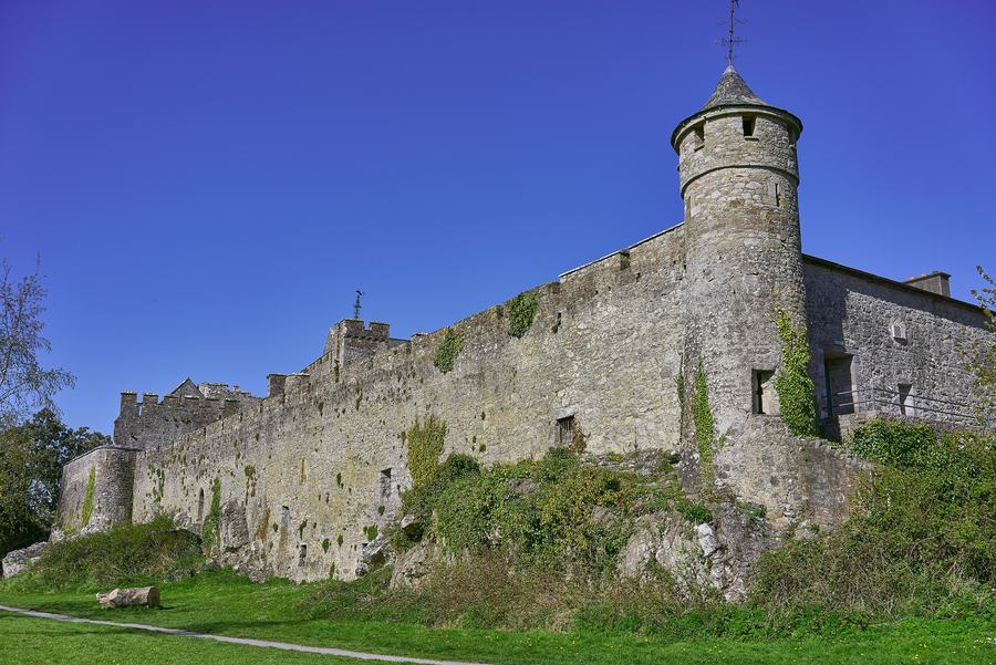 A grey stone castle with a tall round tower and a long battlemented curtain wall rising from a grassy bank under a deep blue sky.