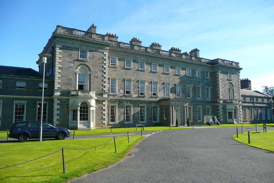 A long two-storey classical house painted pale blue-grey, with a pillared entrance and a gravel forecourt, seen across a lawn.