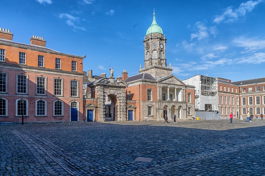 A cobbled courtyard with Georgian ranges in red brick and pale render, a central block with a clock tower and green copper dome, and an arched gateway topped by a statue.