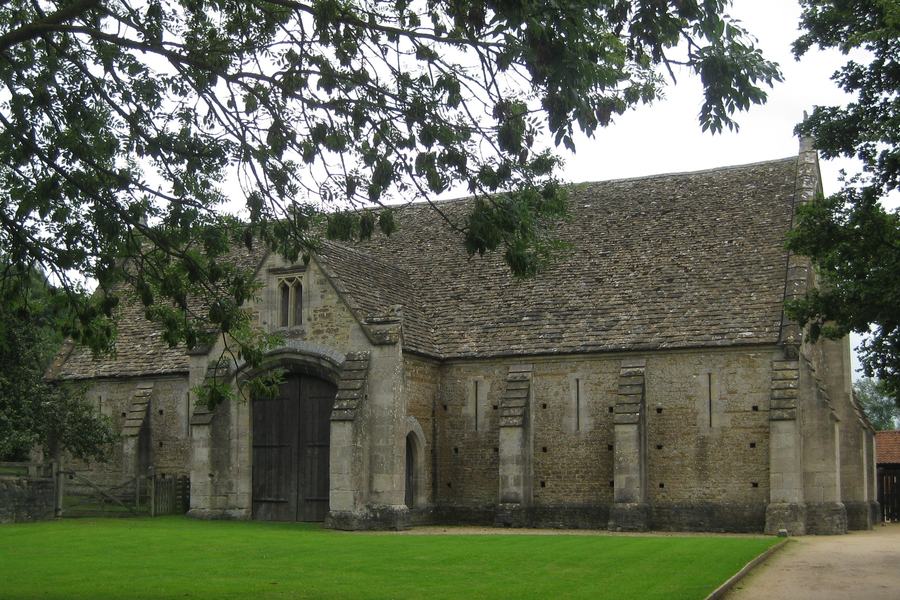 A long medieval stone barn with a steep stone-tiled roof, buttressed walls and a tall gabled porch, standing on mown grass.
