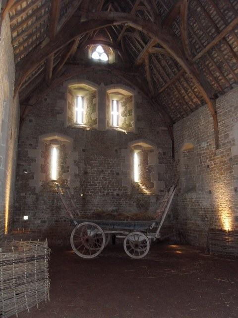 The interior of a medieval barn with a high timber roof of curved trusses, a stone gable pierced by narrow windows, and a wooden farm cart on the earth floor.