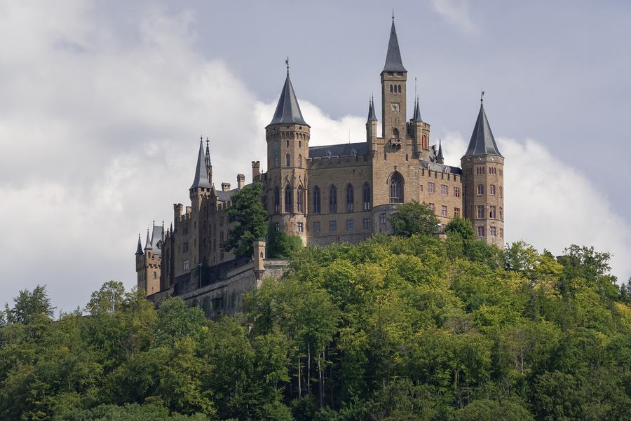 A nineteenth-century castle with many pointed towers and turrets crowning a steep wooded hill above a plain.