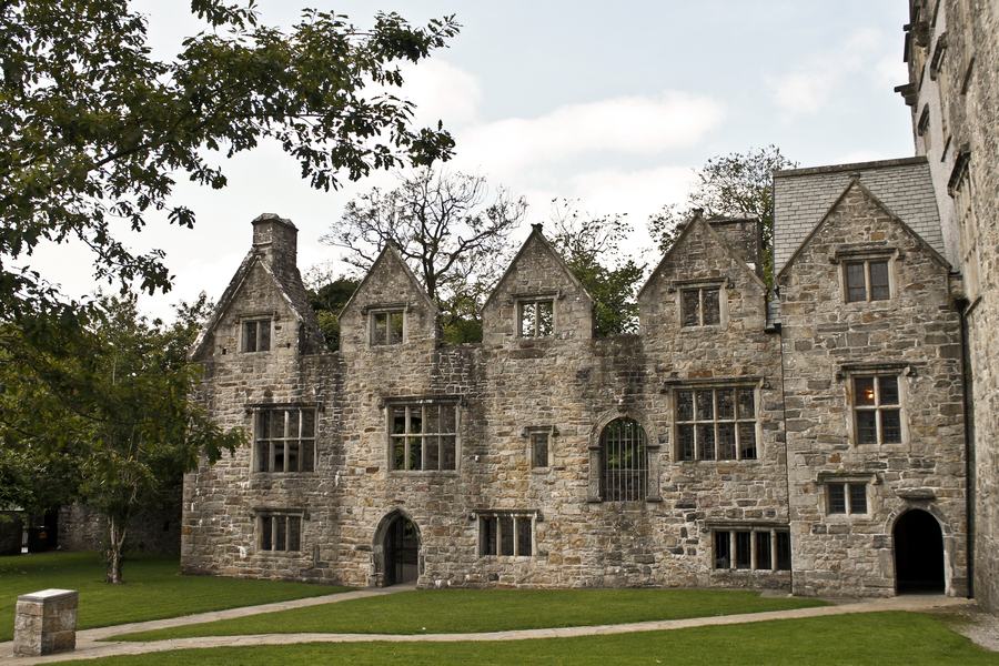 A rambling battlemented country house of grey stone and render, with tall chimneys, seen down a gravel drive past shrubs and lawn.