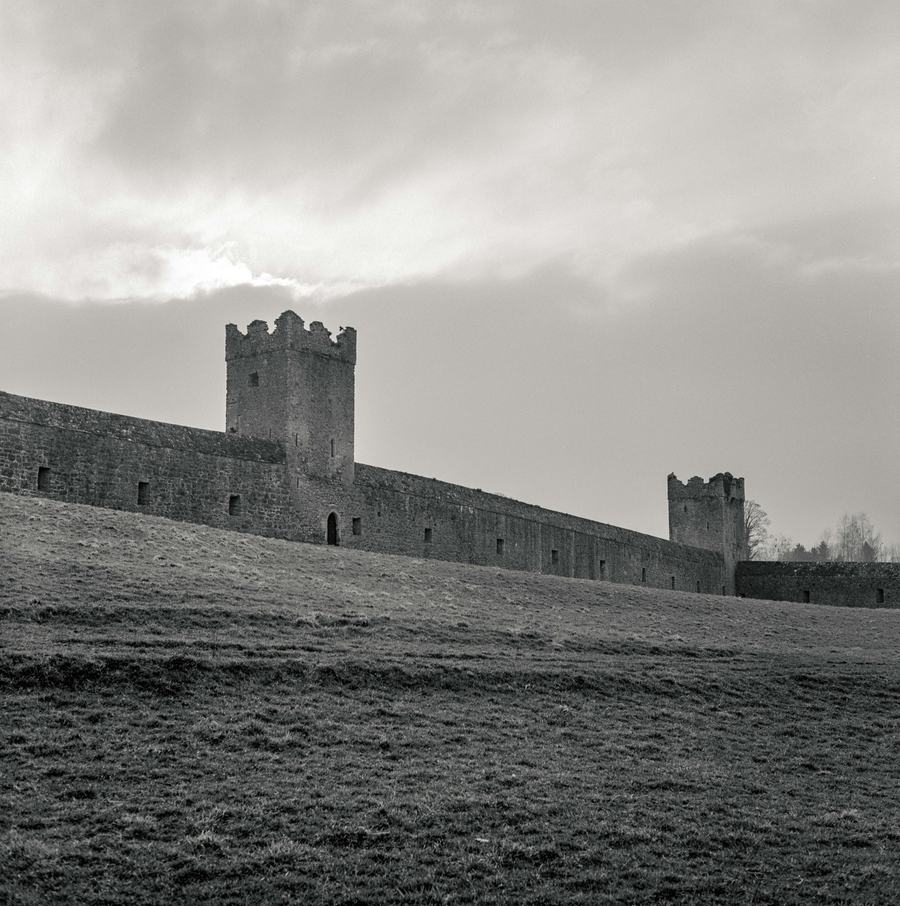 A black-and-white view of a long medieval stone curtain wall with square towers, standing in rough grass under a heavy sky.