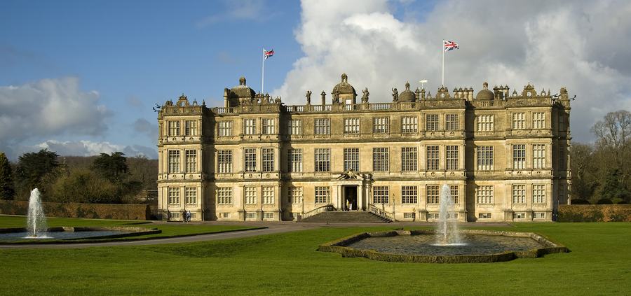 A wide Elizabethan house of pale stone with three storeys of large windows and a flat balustraded roofline, seen across lawns and two fountains.