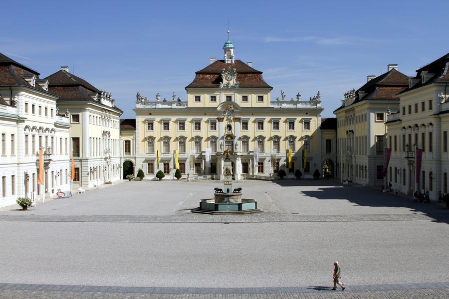A vast baroque palace courtyard enclosed on three sides by pale yellow ranges with mansard roofs, empty except for a single figure crossing the paving.