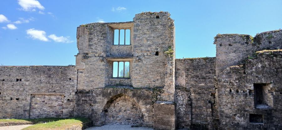 A ruined stone range with a large blocked arch, empty mullioned windows and a square tower at the end, against a clear sky.