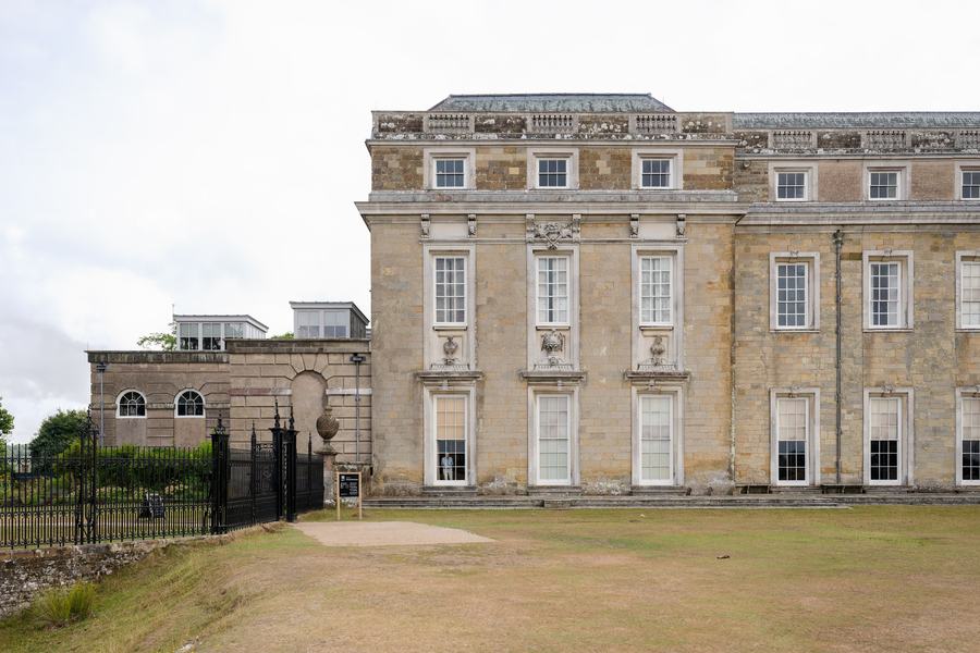 A very long classical house front of pale stone with a regular grid of tall sash windows, standing on open parkland grass.