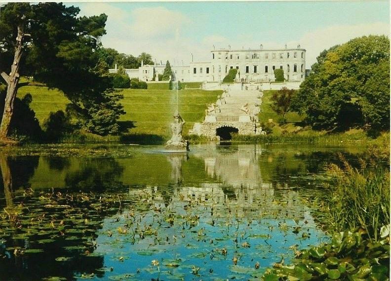 A large classical house on a hillside above a stepped stone cascade and a fountain, reflected in a lily-covered pond framed by trees.