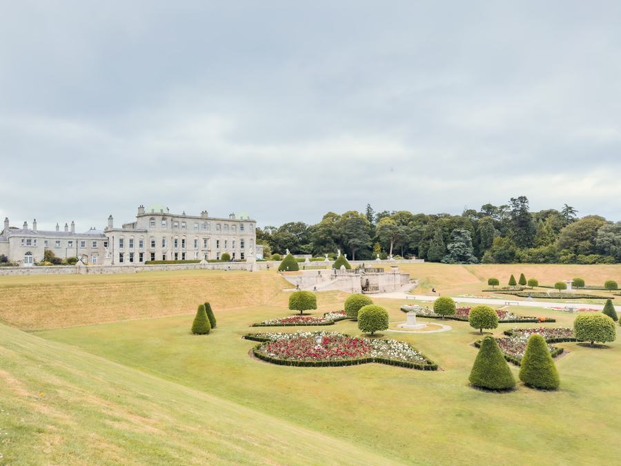 A long pale Georgian house on a ridge above wide mown terraces, with clipped conical yews and a formal flower bed in the foreground under a grey sky.