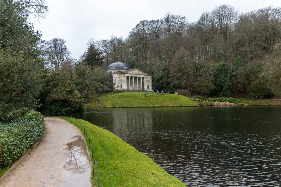 A domed classical temple with a columned portico on the far bank of a still lake, surrounded by bare winter woodland, with a gravel path in the foreground.