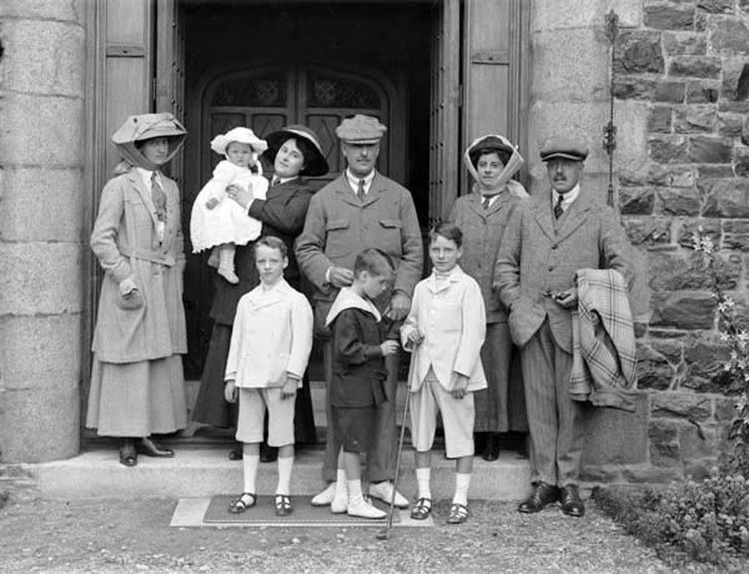 A sepia photograph of a large Edwardian family group, adults and children in white summer dress, posed on the steps of a stone Gothic doorway.