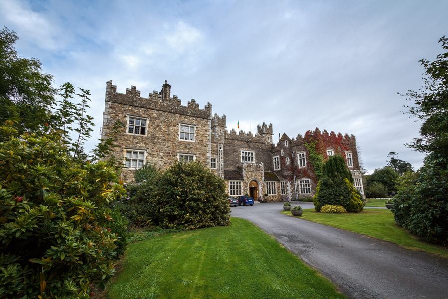 A castellated grey stone house with a square tower and mullioned windows, half hidden among mature trees.