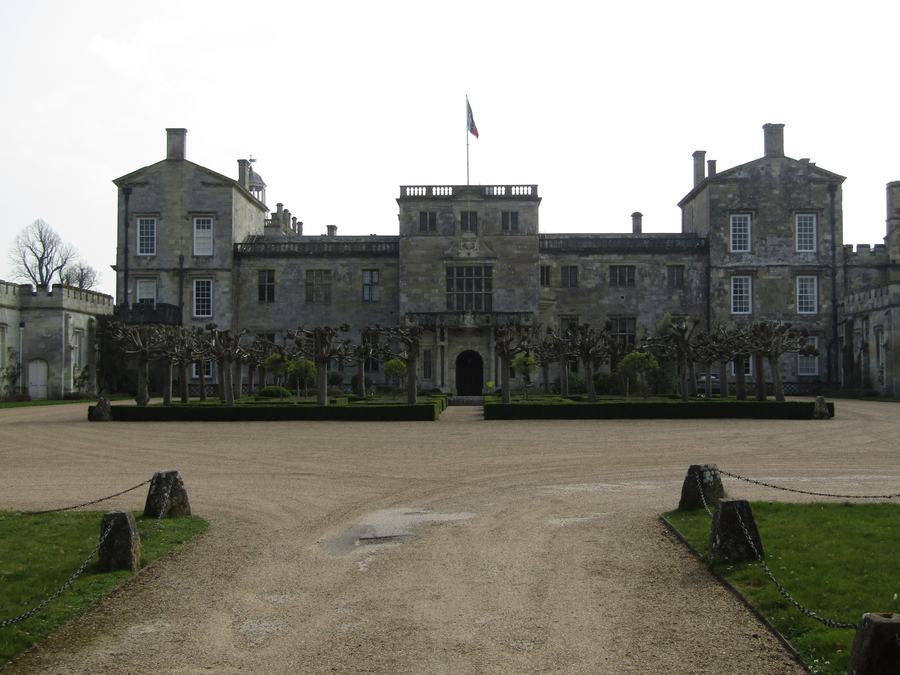 A long two-storey stone house with a central gateway and square corner towers, seen across a gravel forecourt behind iron railings.