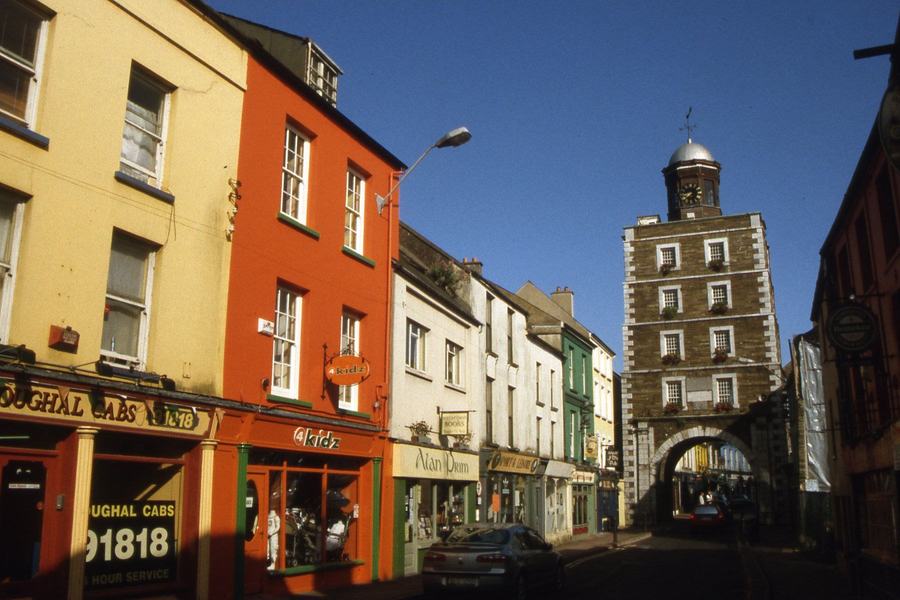 A tall red-and-white eighteenth-century tower with a clock face and lantern, built over an archway across a narrow street of painted shopfronts.