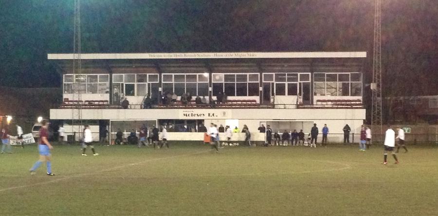 A floodlit evening match at Molesey F.C., with the long clubhouse and its overhanging seating deck along the touchline