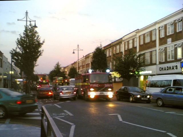Southall Broadway at dusk, shopfronts lit, a fire engine coming down the road