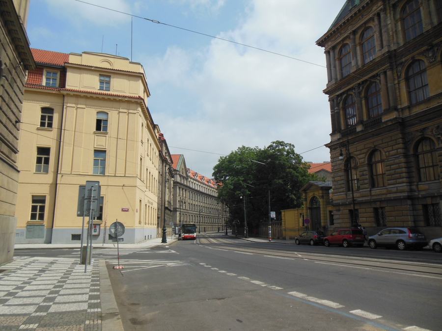 A wide city street with tram tracks, a pale yellow building on the left, an ornate nineteenth-century museum front on the right, a red tram in the distance, summer trees at the end of the street.