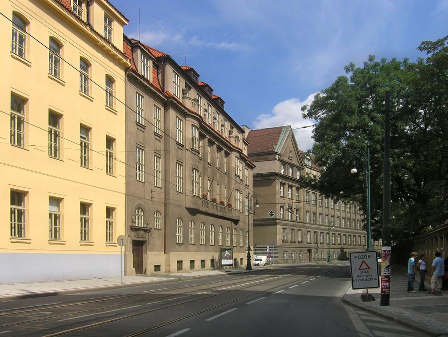 A street with tram rails in the roadway, tall plain buildings on the left, a long wall and mature trees on the right, a few pedestrians and a warning sign on the pavement.