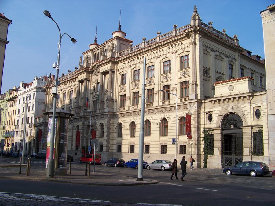 A long ornate nineteenth-century palace front on a city square, two storeys with pilasters and sculpted figures on the roofline, lettering reading BOSCOLO LUXURY HOTEL, cars and pedestrians in front.