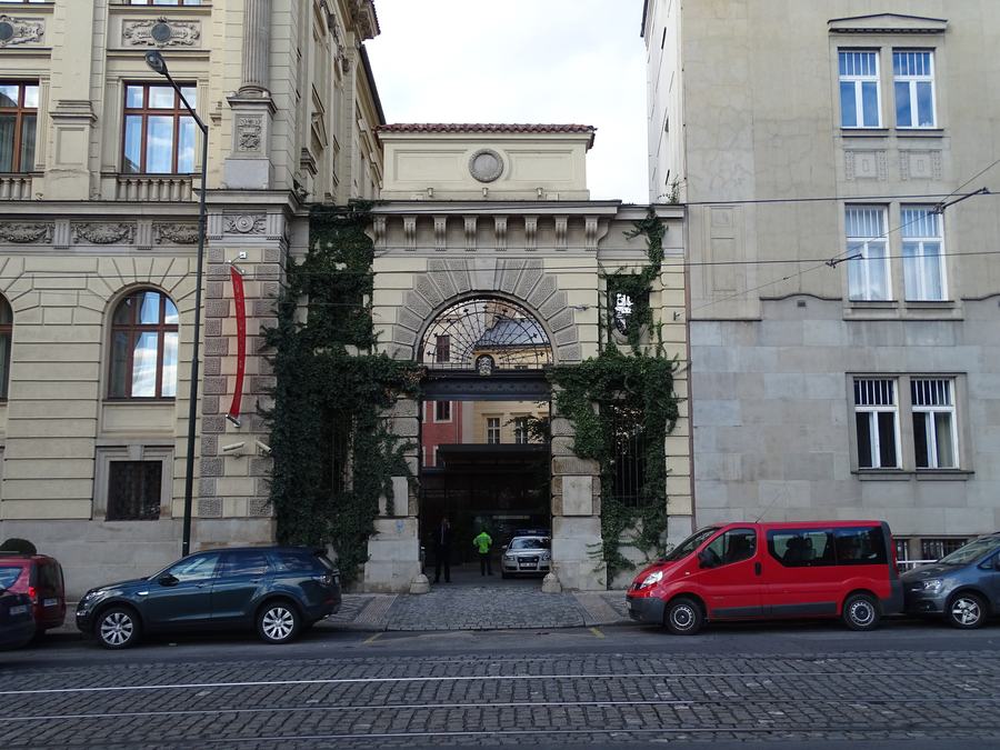 A tall arched gateway with a fanlight of ironwork set in a stone frame, ivy growing up both sides, a passage and a lit doorway visible through it, tram rails and parked cars in front.