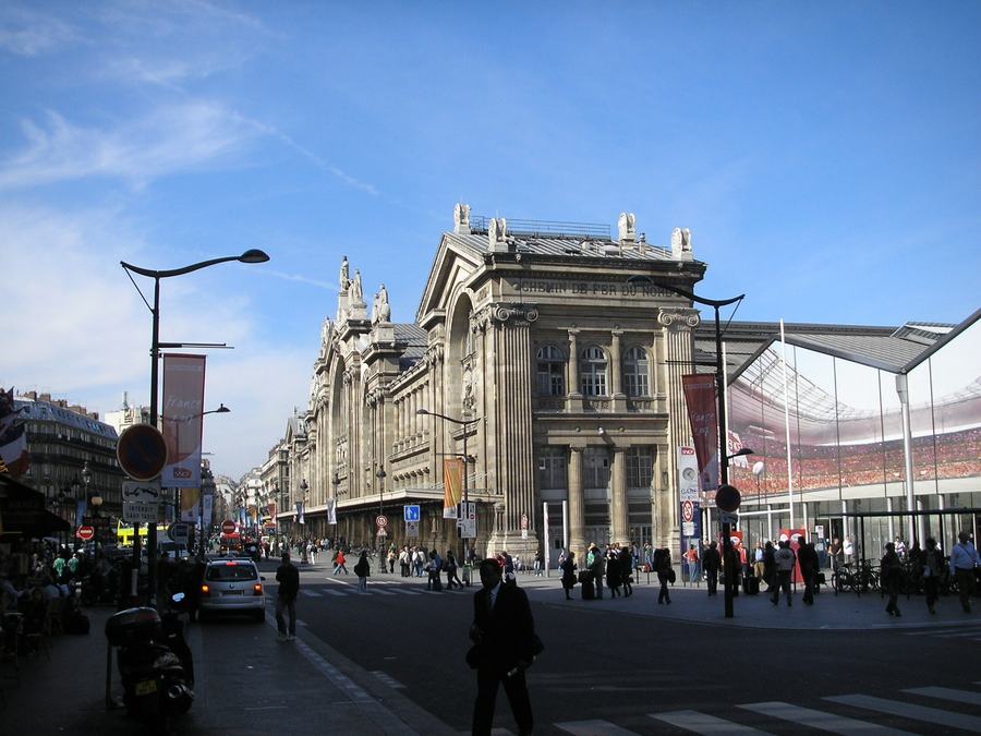 The stone facade of a large railway station with a huge arched window, columns and statues along the roofline, a modern glass canopy to the right, traffic and pedestrians on the street in front.