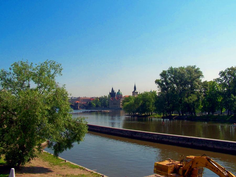 A grassy riverbank with willows on the left, the Vltava with a weir running across the frame, bridge towers and church domes on the far bank under a clear blue sky.