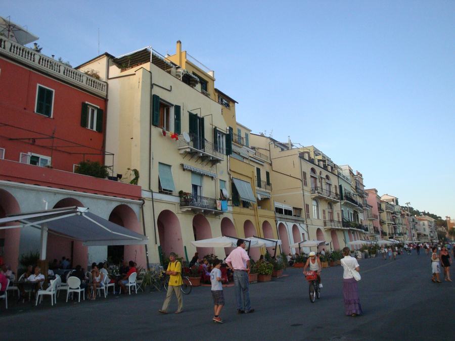 A row of tall coloured houses with arcades at ground level along a harbour promenade, cafe tables and white parasols under the arches, people walking and cycling on the open quay in evening light.