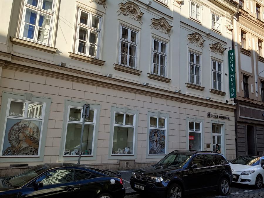 A pale baroque-revival facade on a narrow street, first-floor windows with carved pediments, a green vertical sign reading MUCHA MUSEUM, Art Nouveau posters in the ground-floor windows, cars parked at the kerb.
