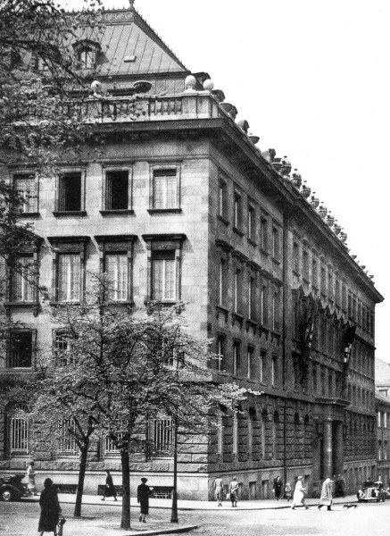 Black-and-white photograph of the same stone palace corner, a large swastika banner hanging from the upper floor, bare trees and pedestrians in period coats on the pavement, a 1930s car at the kerb.