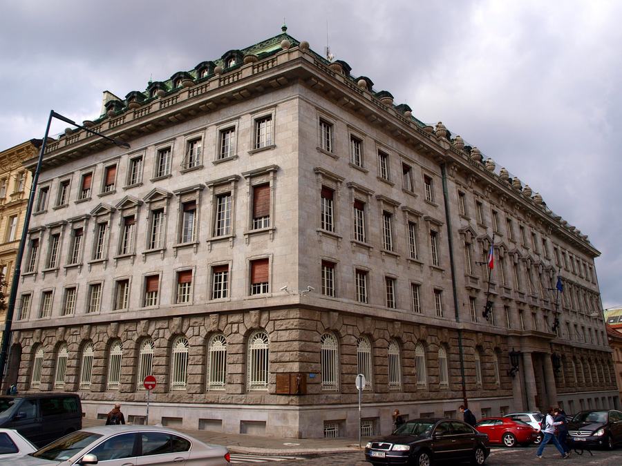 A four-storey neo-classical stone palace on a street corner, rusticated ground floor with arched barred windows, rows of tall rectangular windows above, a green copper mansard roof, a Czech flag over the entrance on the right.
